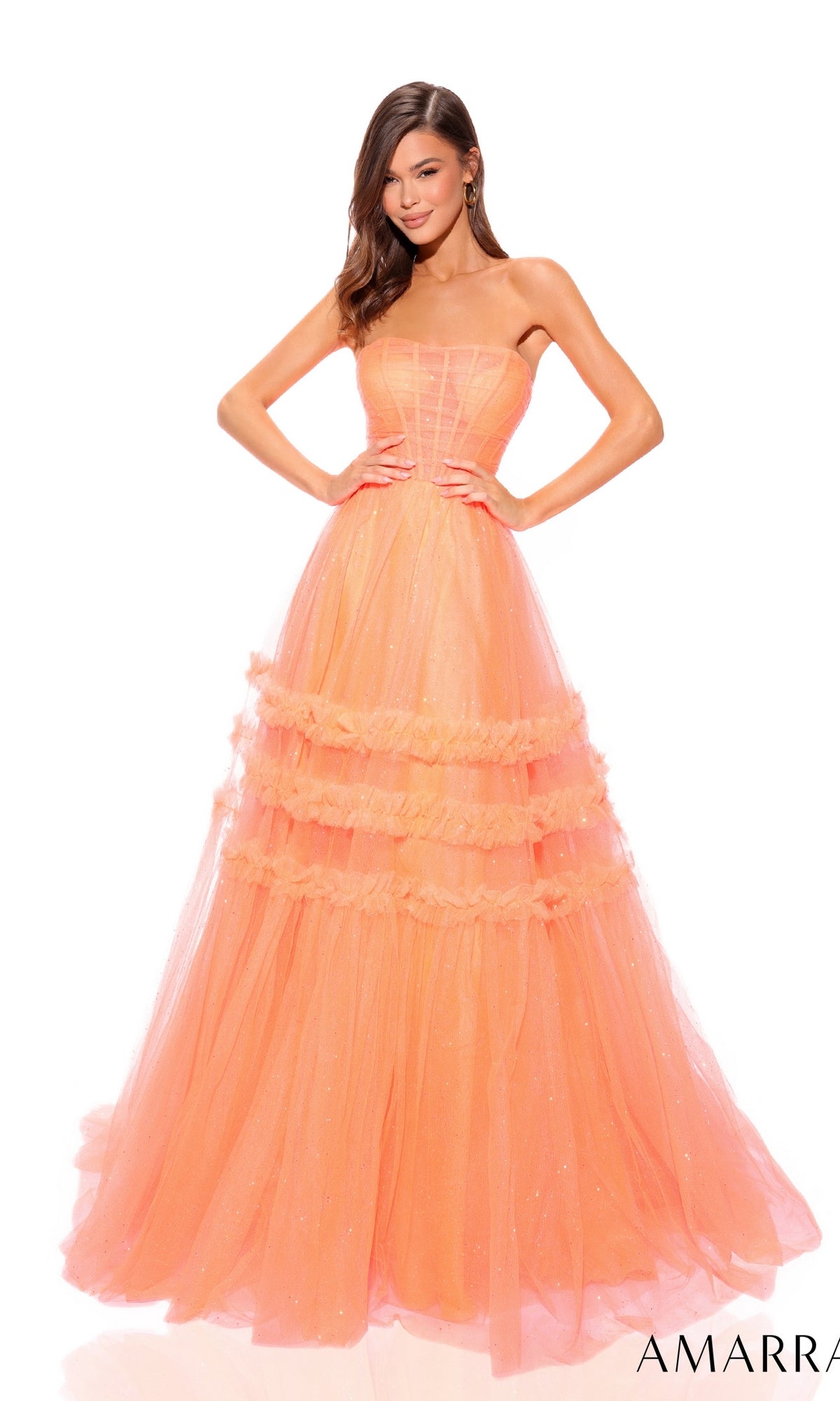 A woman models the Amarra Strapless Sheer-Bodice Prom Ball Gown 88794, featuring ruffled layers and a peach hue, as she poses with her hands on her hips against a white background.